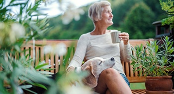 person drinking a cup of coffee and petting their dog while sitting on a bench in their backyard